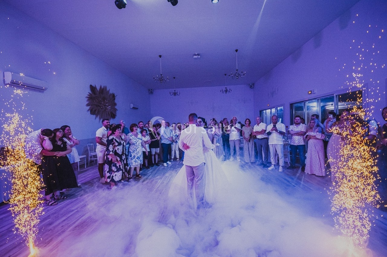 Un couple danse au centre d'une salle entourée d'invités, avec des étincelles et de la fumée au sol.