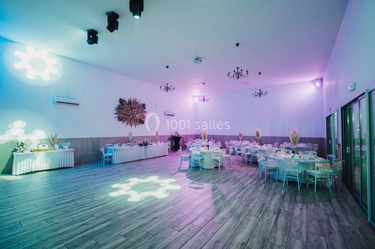 Salle de réception décorée avec des tables rondes dressées, éclairage tamisé et projecteurs lumineux au plafond.