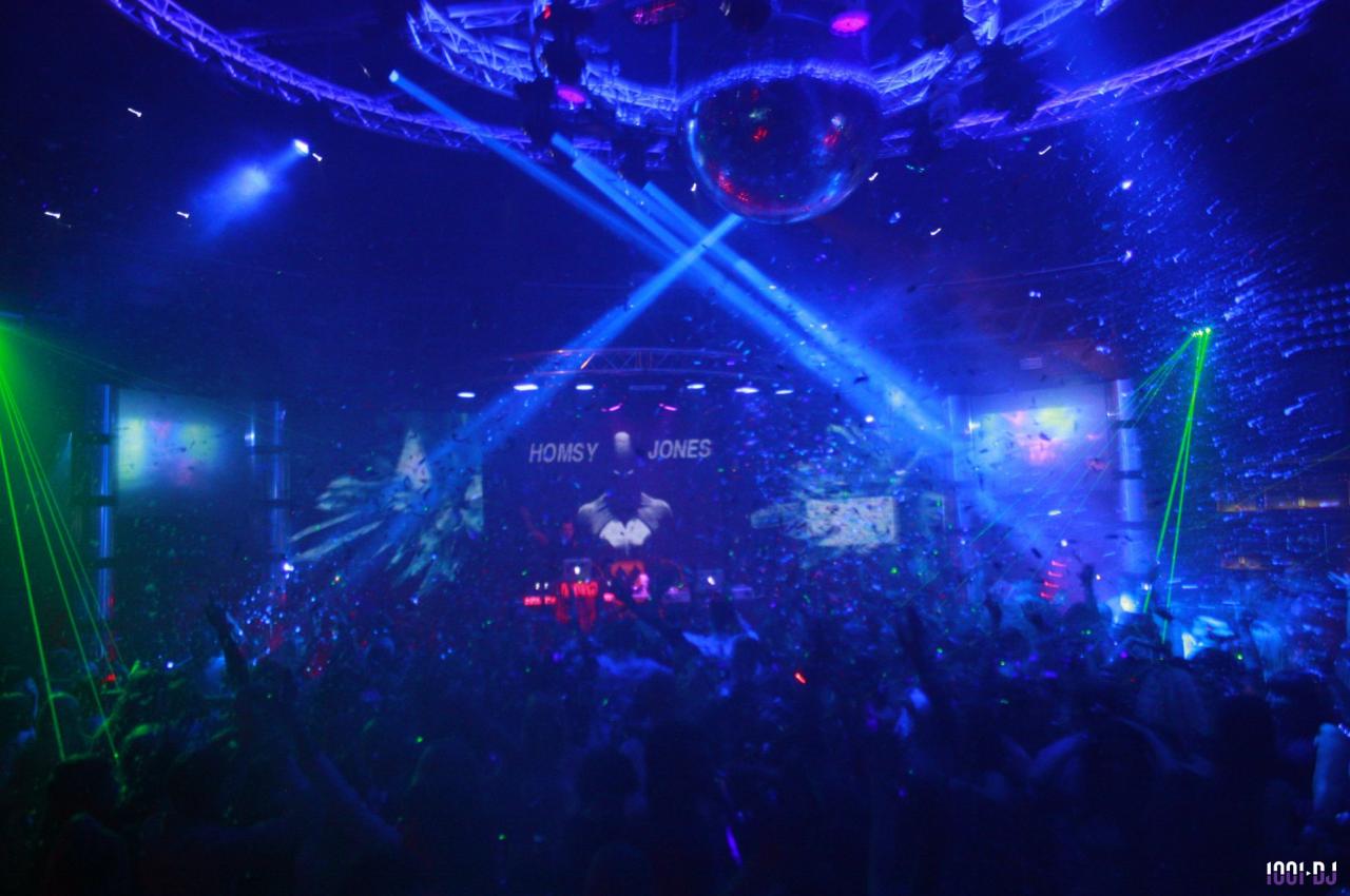 Foule dans une discothèque sombre avec des jeux de lumières colorées, une boule disco et une scène illuminée.