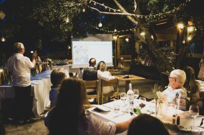 Des personnes assises à l'extérieur, regardant une présentation projetée sur un écran sous des guirlandes lumineuses.