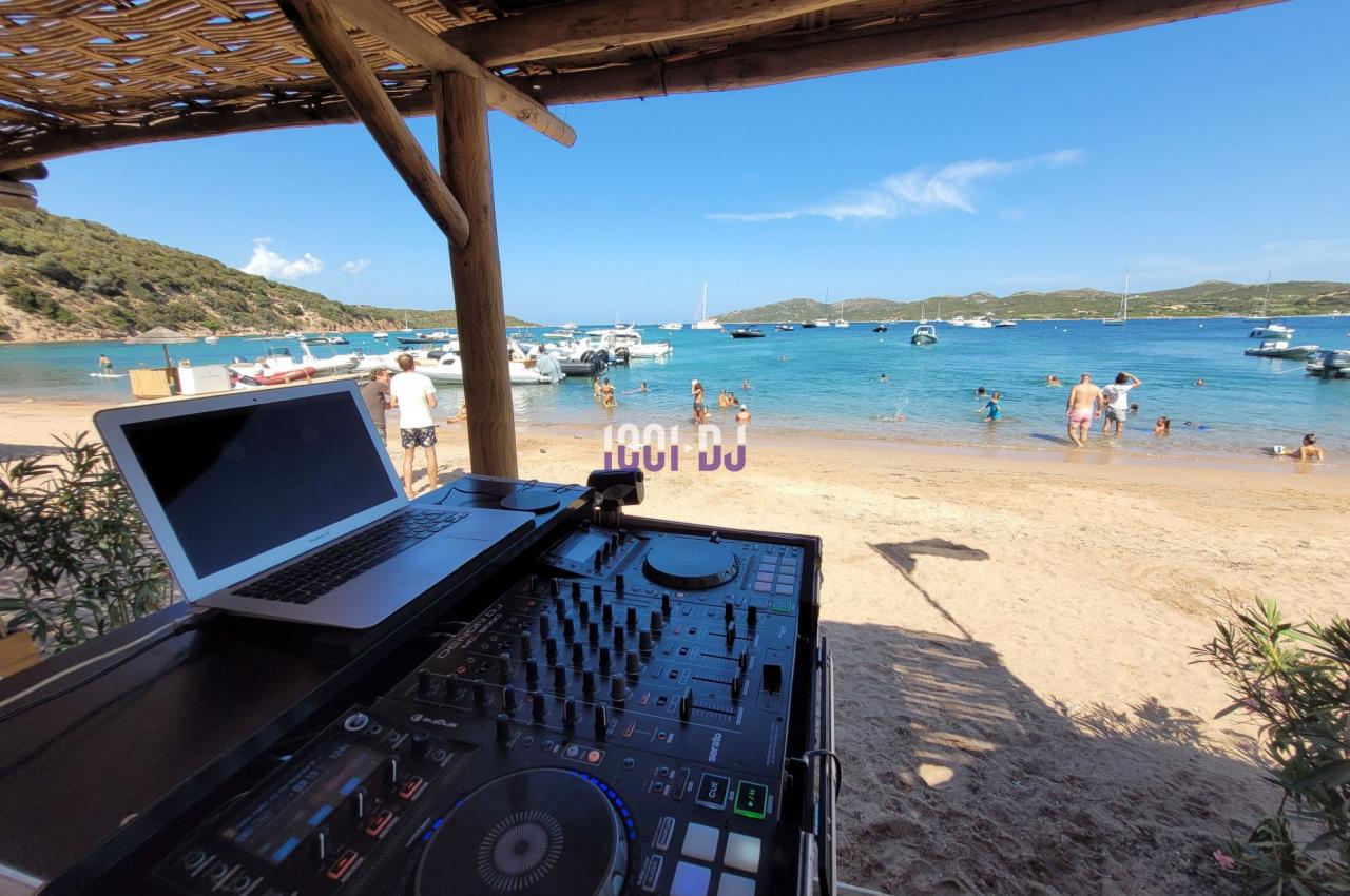 Table de mixage et ordinateur portable installés sous un abri en bois, avec vue sur une plage et des bateaux amarrés.