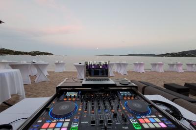 Table de mixage DJ sur une plage au coucher du soleil, avec des tables dressées en arrière-plan près de la mer.