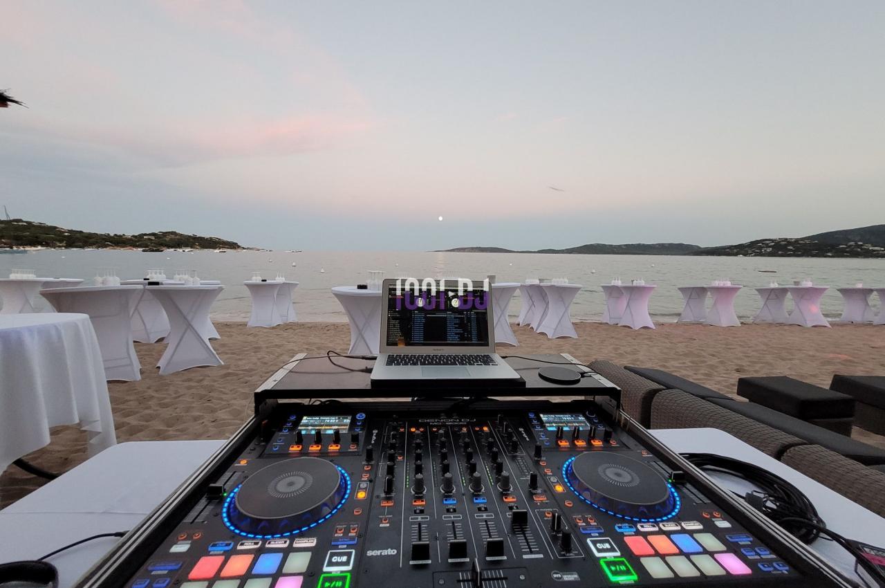 Table de mixage DJ sur une plage au coucher du soleil, avec des tables dressées en arrière-plan près de la mer.