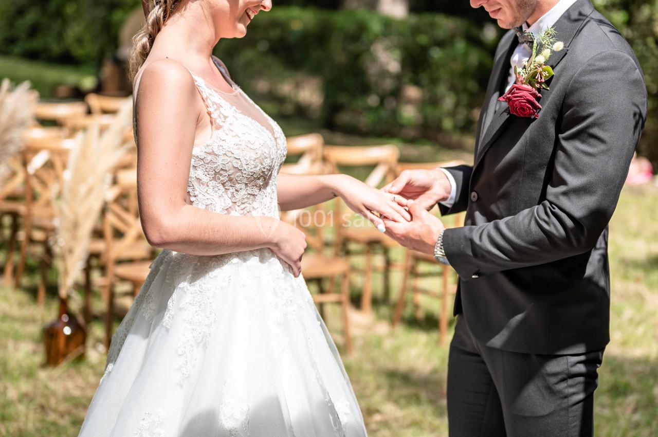 Un couple échange des alliances lors d'une cérémonie de mariage en extérieur, entouré de verdure et de chaises en bois.