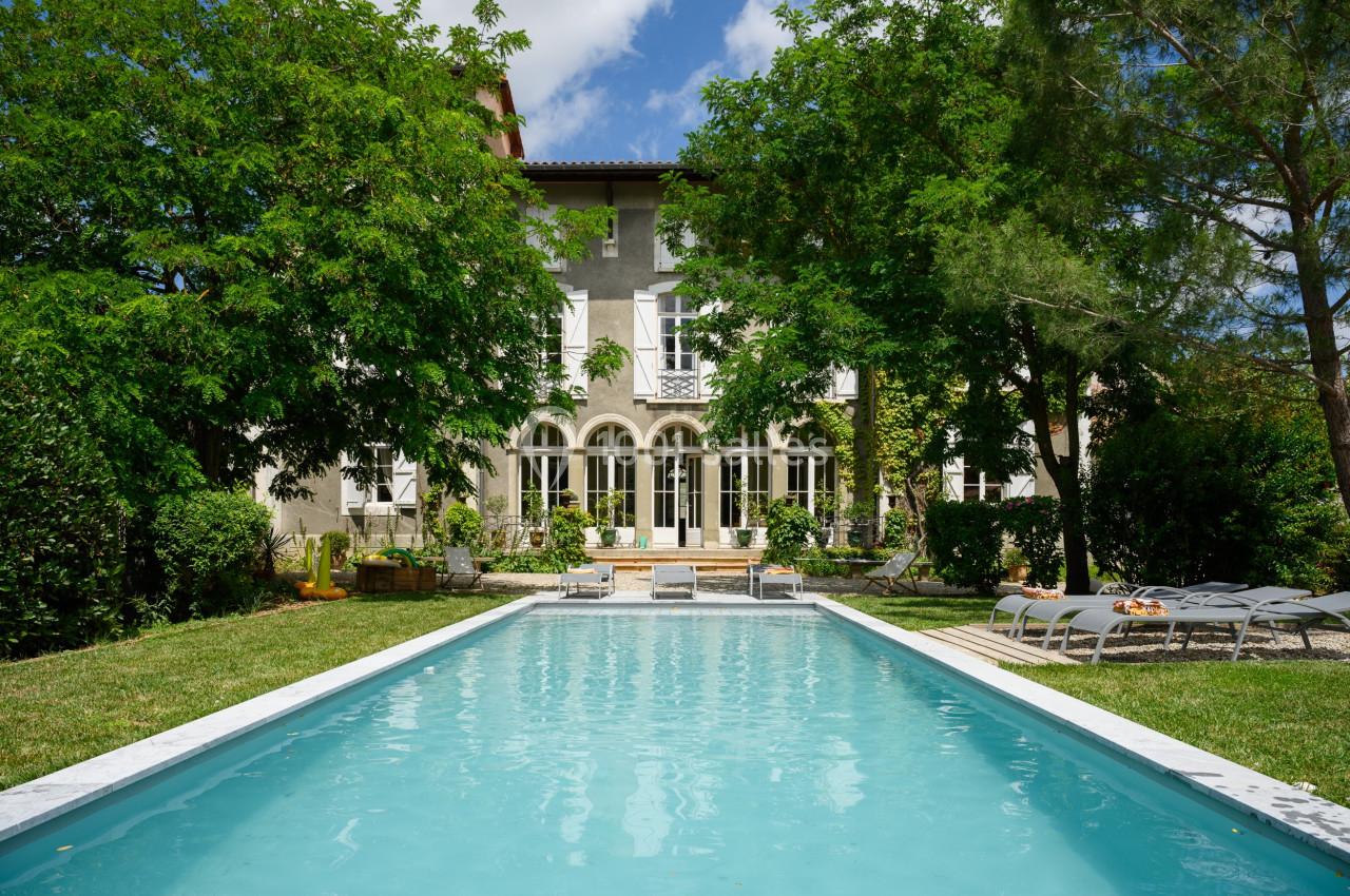 Grande maison entourée d'arbres avec une piscine rectangulaire au premier plan et des chaises longues sur le côté.