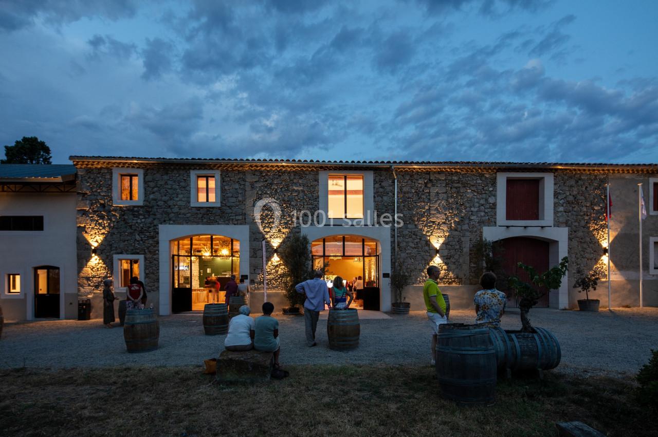 Façade en pierre d'un bâtiment éclairé au crépuscule, avec des personnes assises et debout devant des tonneaux.