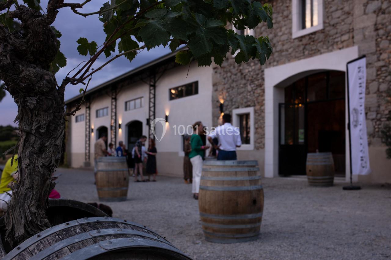Des personnes discutent près de tonneaux en bois devant un bâtiment en pierre, dans une ambiance de fin de journée.