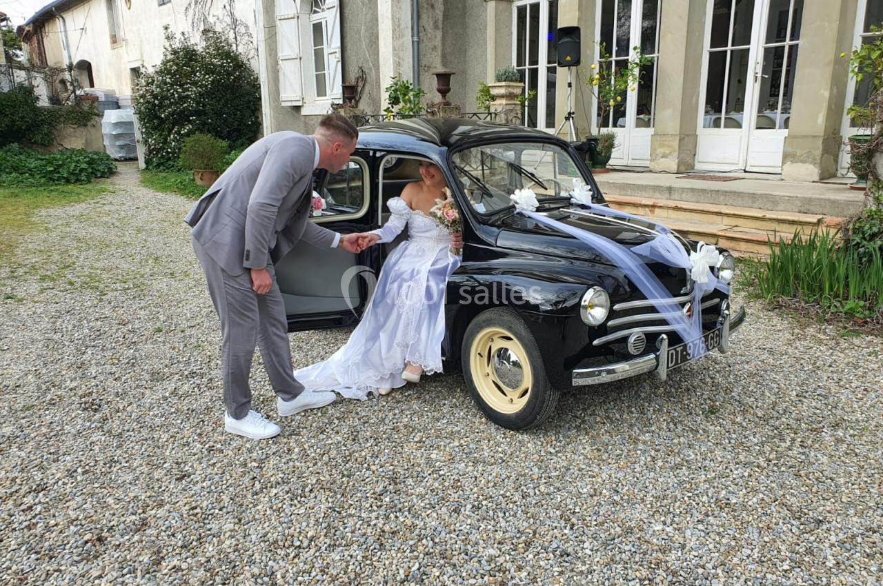 Un marié aide sa partenaire en robe de mariée à sortir d'une voiture ancienne décorée pour un mariage.