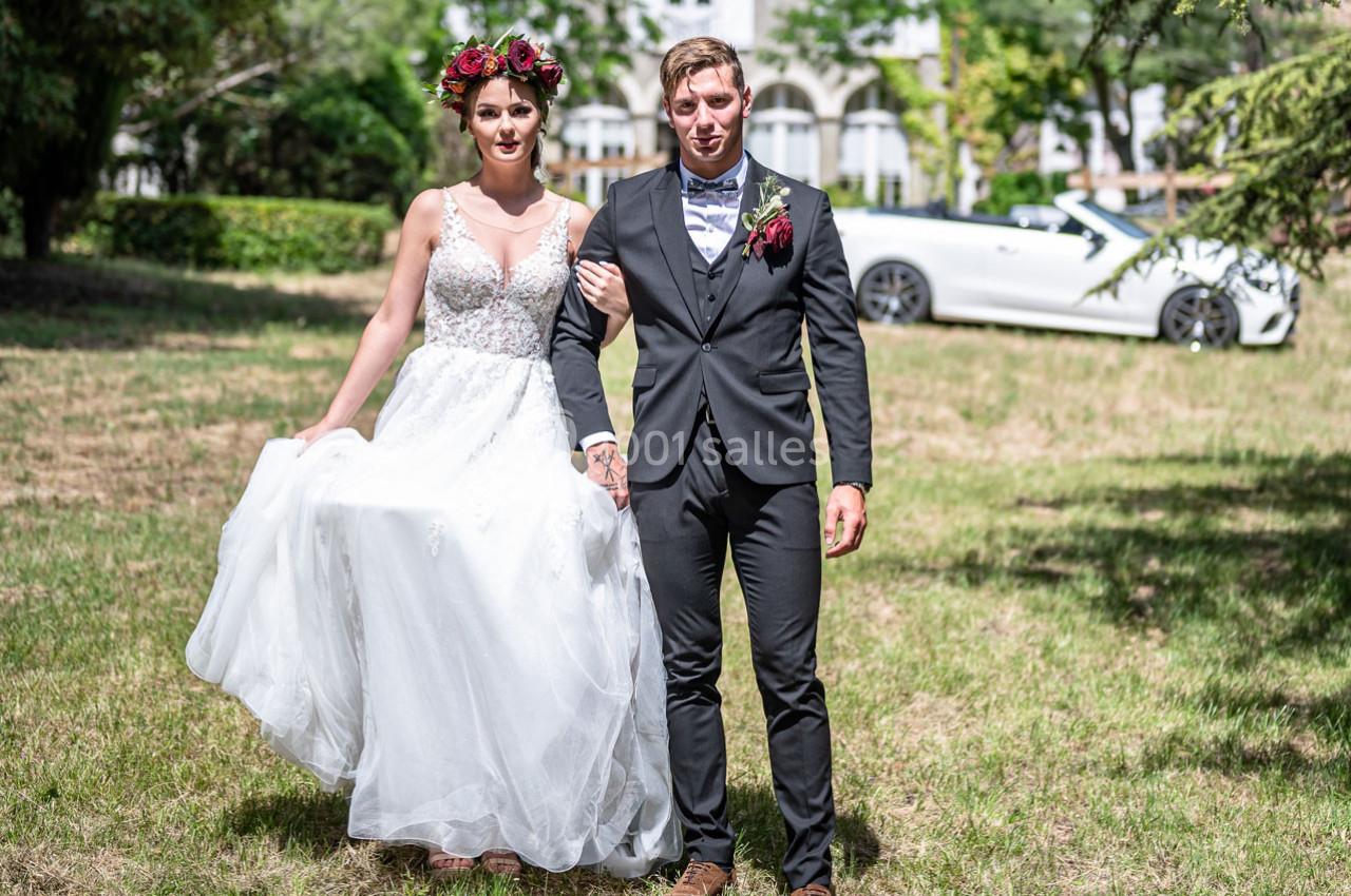Un couple en tenue de mariage marche sur une pelouse, avec une voiture blanche décapotable en arrière-plan.