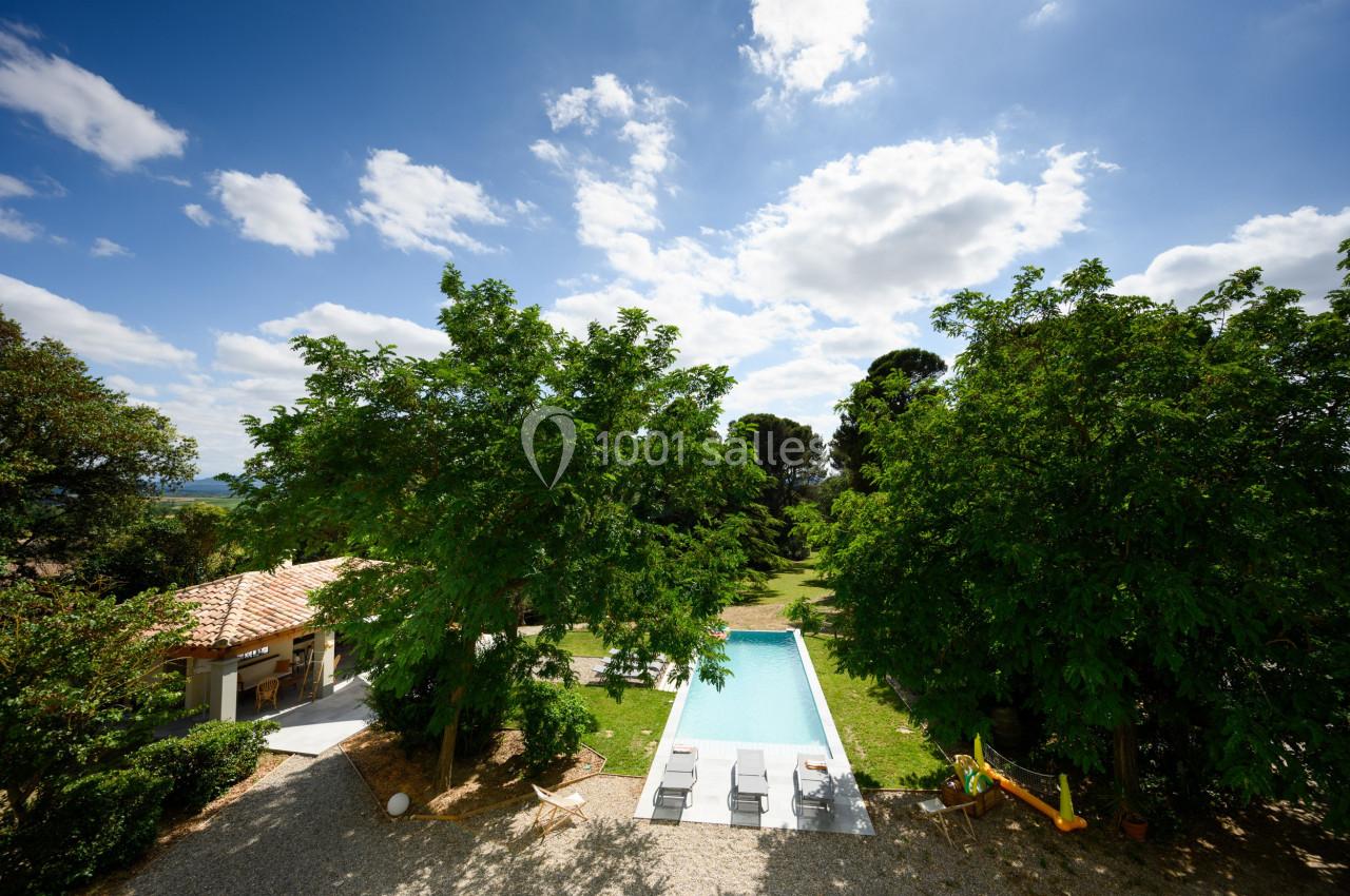Vue aérienne d'une piscine entourée de verdure, avec des chaises longues et un bâtiment annexe sous un ciel dégagé.