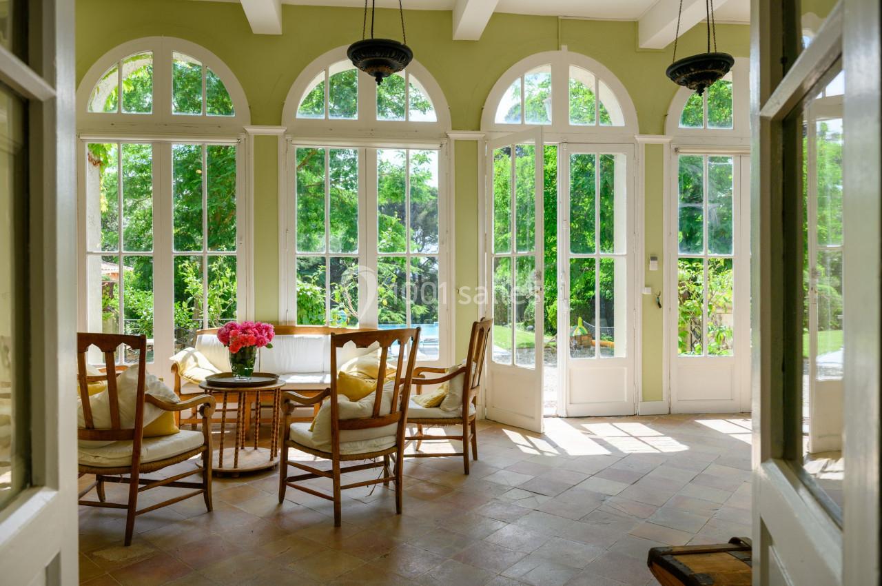 Salle lumineuse avec de grandes baies vitrées, des chaises en bois autour d'une table et vue sur un jardin verdoyant.