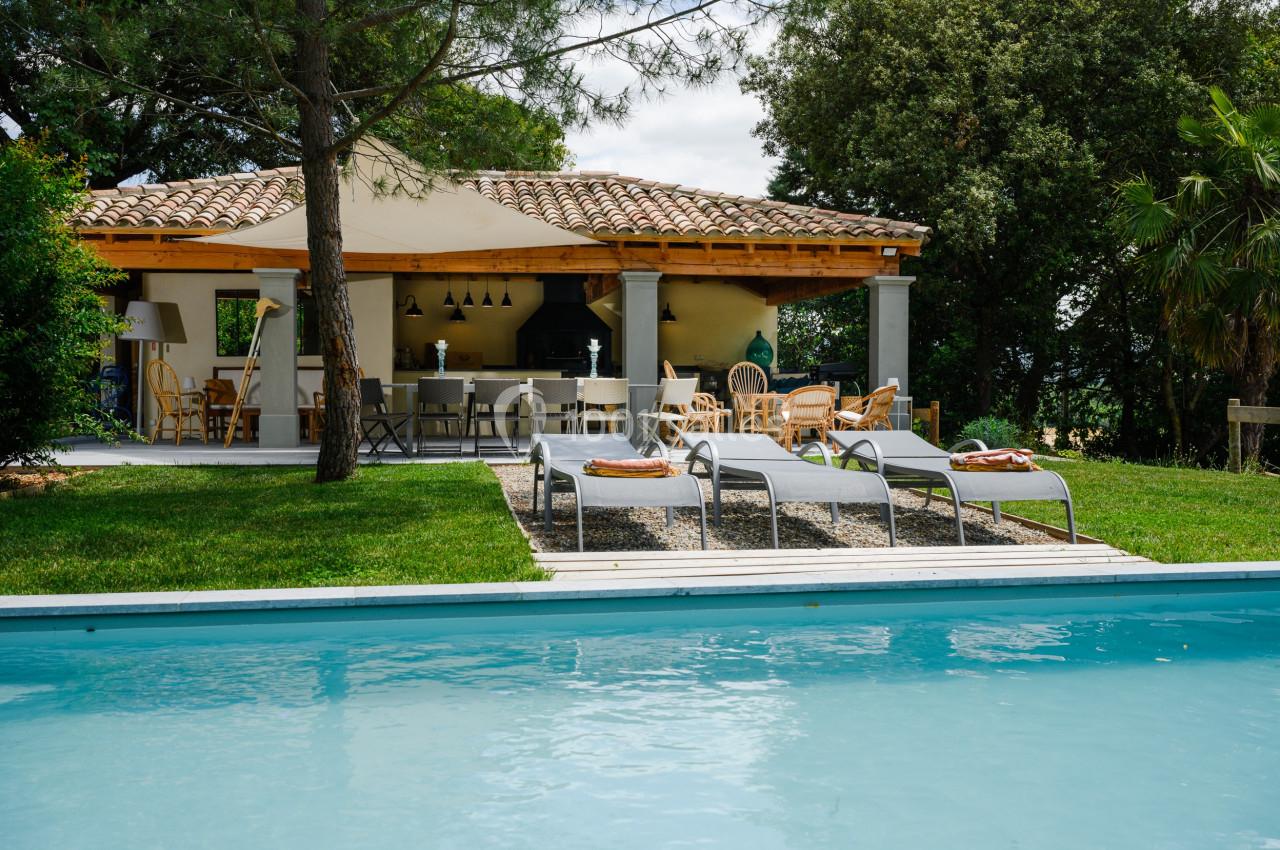 Terrasse avec mobilier de jardin sous une pergola, vue sur une piscine entourée de pelouse et d'arbres.