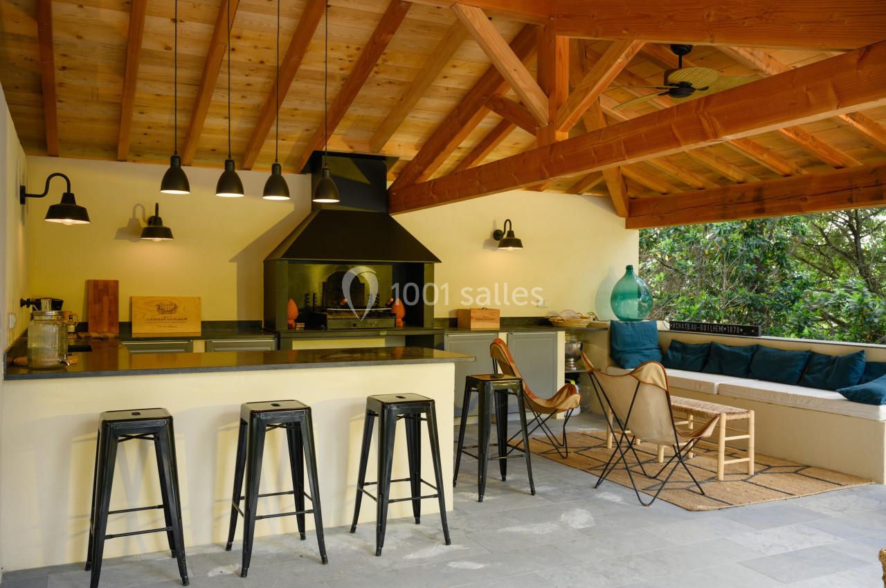 Cuisine d'extérieur couverte avec toit en bois, comptoir avec tabourets, barbecue, et coin salon avec canapé.