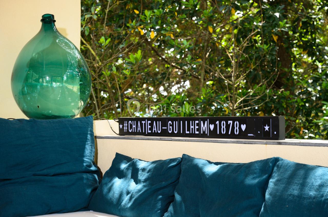 Coussin bleu sur un banc extérieur avec une carafe en verre vert et une pancarte décorative ’Château Guilhem 1878’.