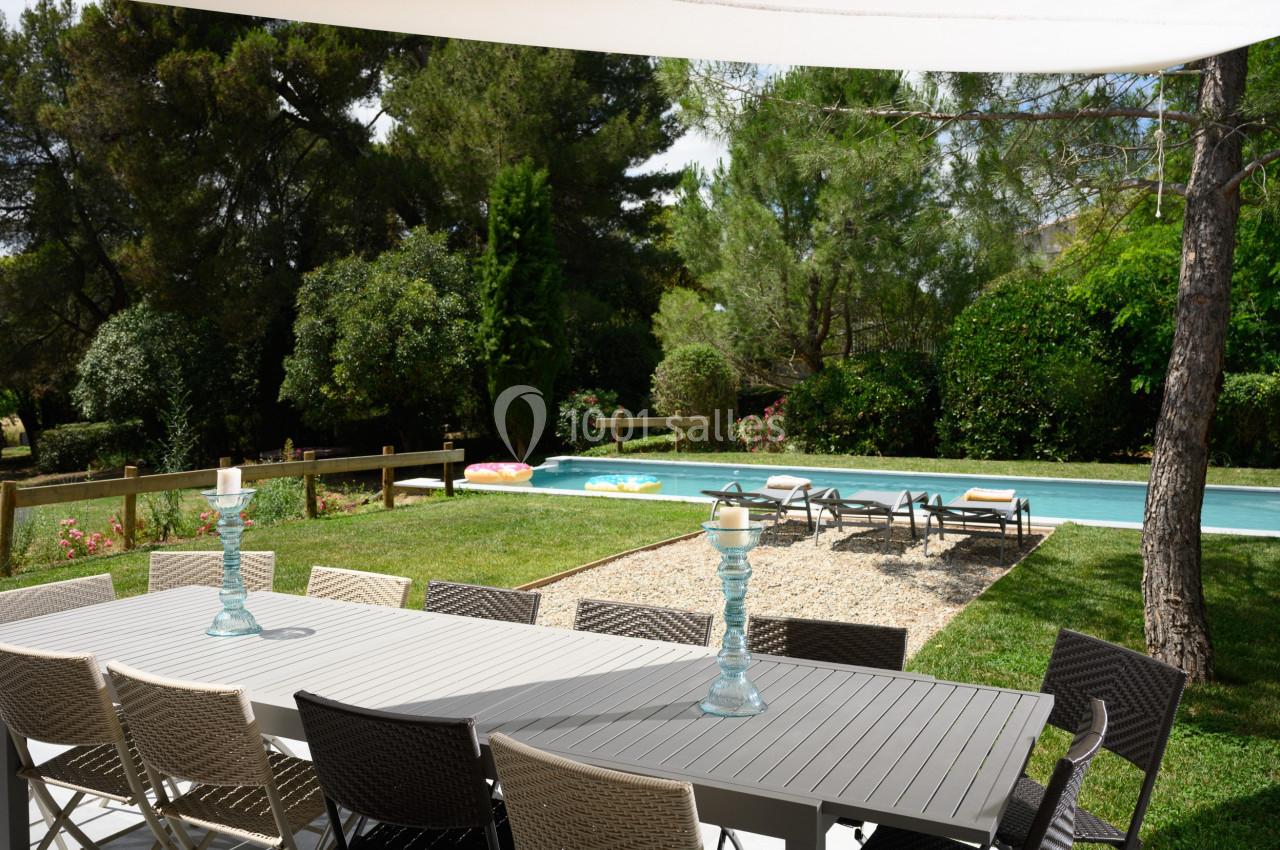 Table de jardin avec chaises, vue sur une piscine entourée de transats, pelouse et arbres en arrière-plan.