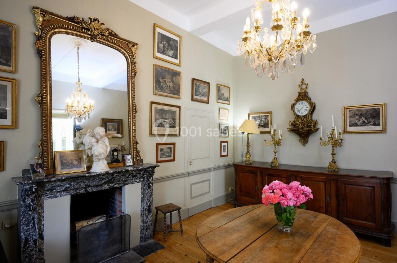 Salle élégante avec cheminée en marbre, miroir doré, lustres, meubles anciens et bouquet de fleurs sur une table en bois.