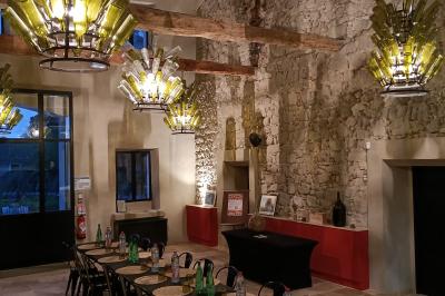 Salle en pierre avec de grandes tables dressées, éclairée par des lustres faits de bouteilles en verre vert.