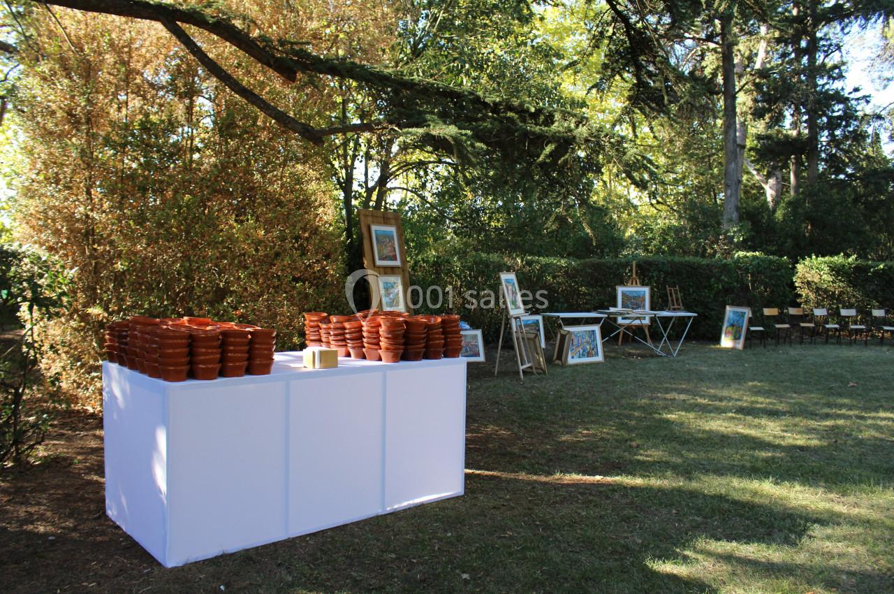 Table blanche avec des pots en terre cuite empilés, entourée de cadres et de chaises dans un jardin arboré.