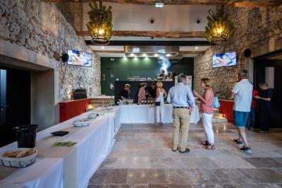 Salle en pierre avec de grandes tables dressées, éclairée par des lustres faits de bouteilles en verre vert.