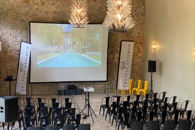 Salle en pierre avec de grandes tables dressées, éclairée par des lustres faits de bouteilles en verre vert.