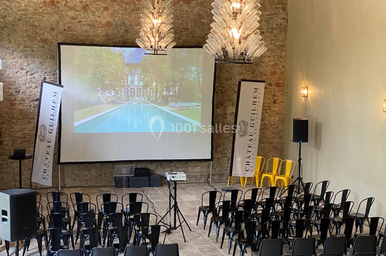 Salle de conférence avec des rangées de chaises, un écran de projection allumé et des bannières promotionnelles.