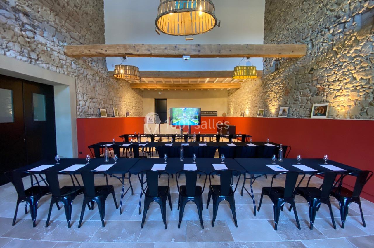 Salle de réunion avec murs en pierre, tables disposées en U, chaises noires et écran de présentation au fond.