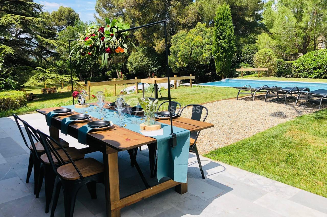 Table en bois dressée avec vaisselle et serviettes bleues, installée sur une terrasse près d'une piscine et d'un jardin…
