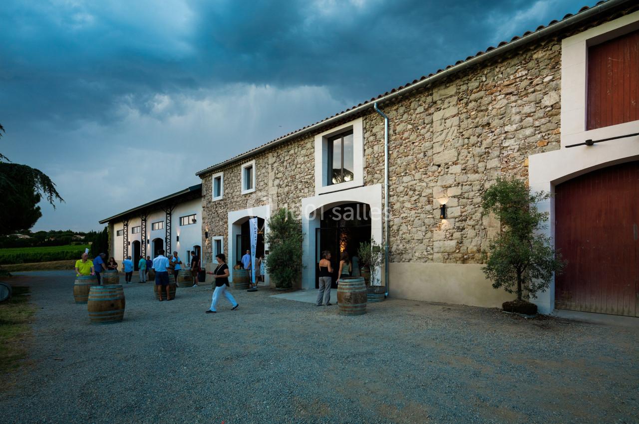 Façade en pierre d'un domaine viticole avec des visiteurs entrant et sortant sous un ciel nuageux.