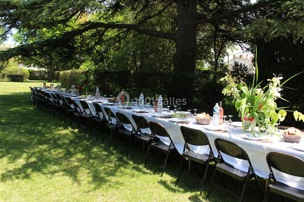 Table longue dressée en extérieur sous des arbres, avec nappes blanches, chaises noires et décoration florale.