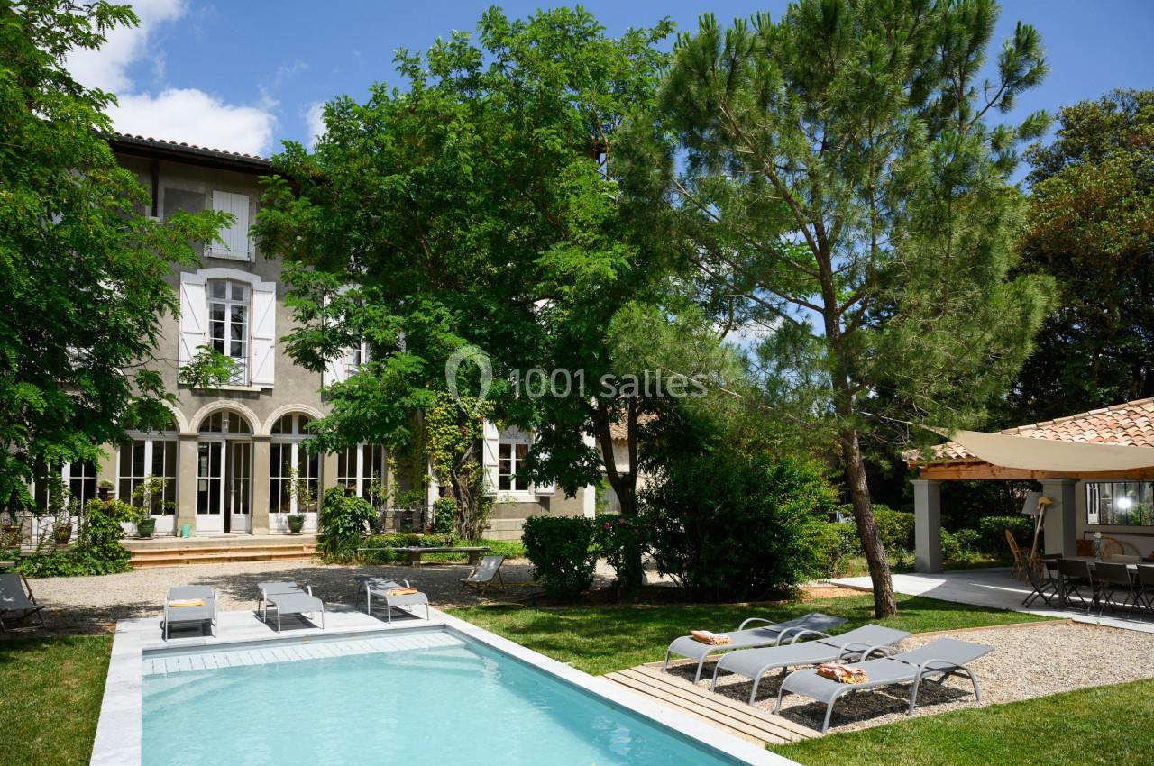Maison avec façade claire, jardin arboré, piscine entourée de transats et terrasse ombragée sous un ciel dégagé.