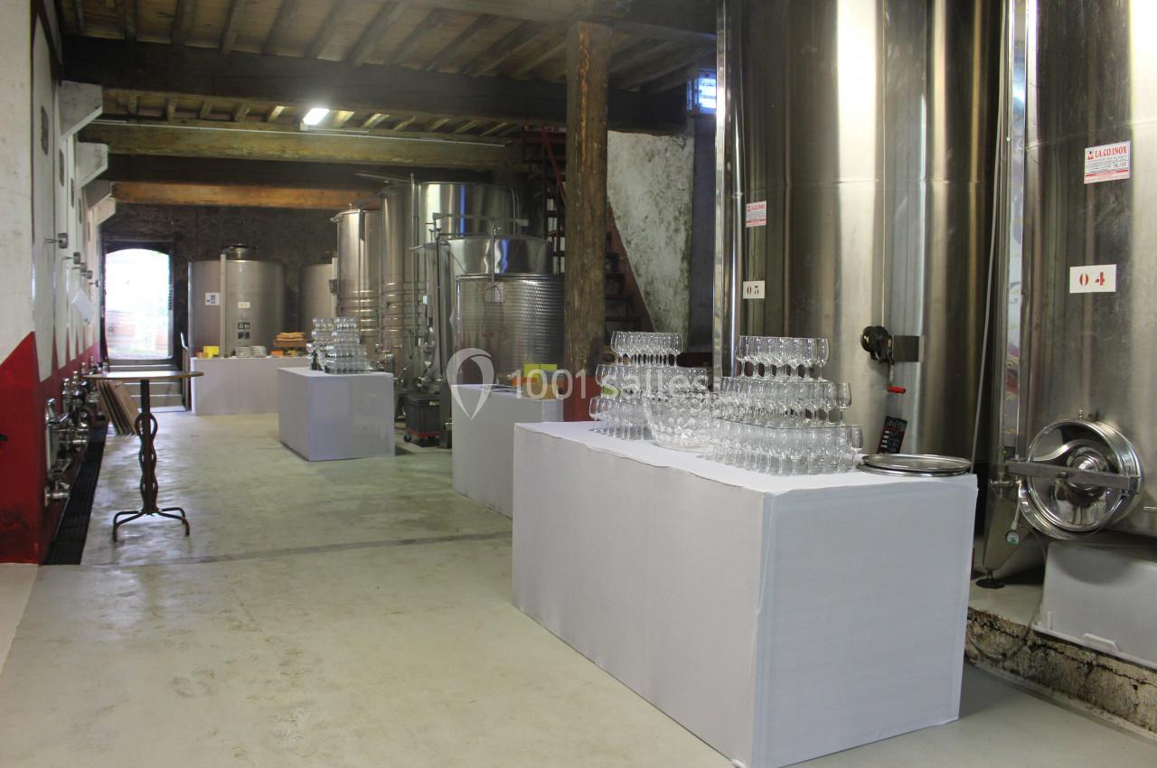 Salle de stockage avec cuves en inox, tables couvertes de nappes blanches et verres empilés pour une dégustation.