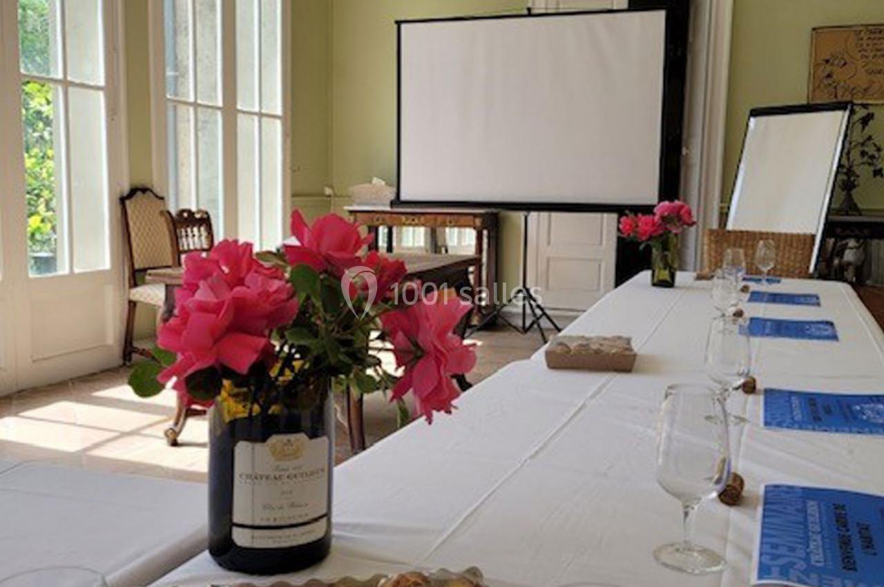 Salle lumineuse avec table dressée, fleurs roses en vase, écran de projection et documents disposés.