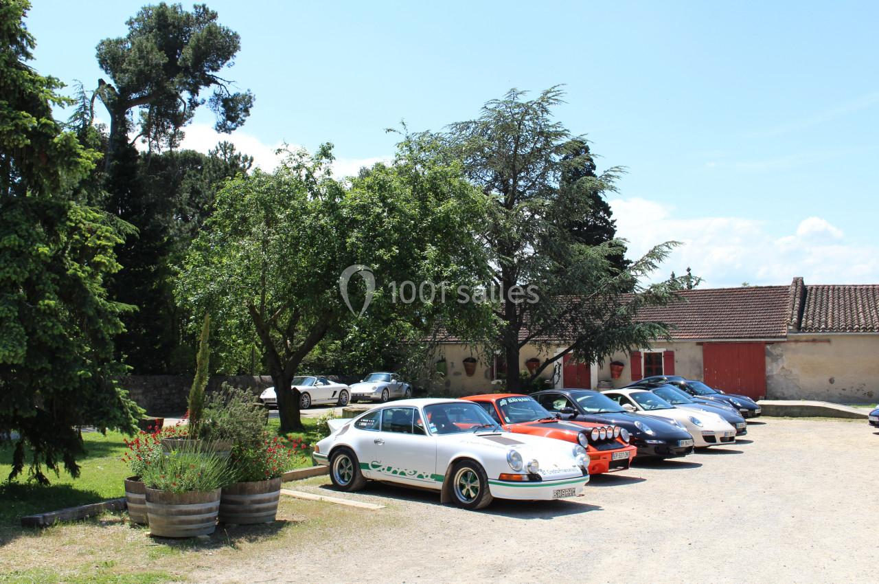 Plusieurs voitures anciennes stationnées dans une cour ensoleillée, entourée d'arbres et de bâtiments rustiques.