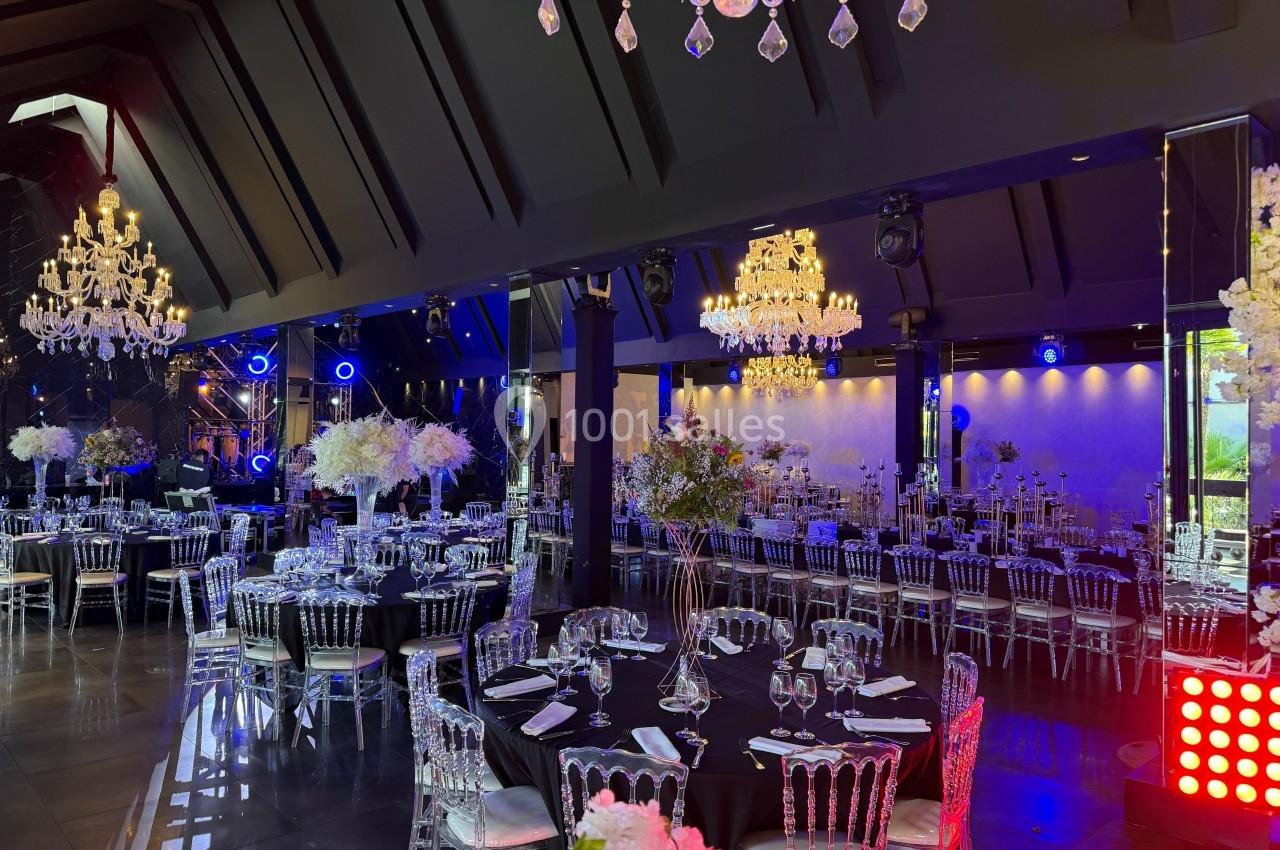 Salle de réception élégante avec tables dressées, chaises transparentes, lustres en cristal et éclairage d'ambiance.