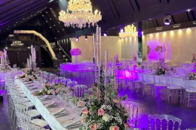 Salle de réception élégante avec tables rondes dressées, chaises transparentes et lustres suspendus.