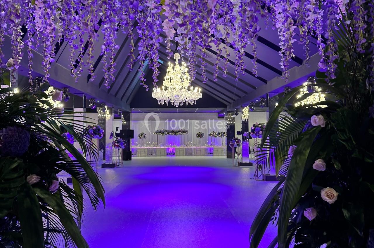 Salle de réception illuminée en violet, décorée de guirlandes florales suspendues et d'un grand lustre central.