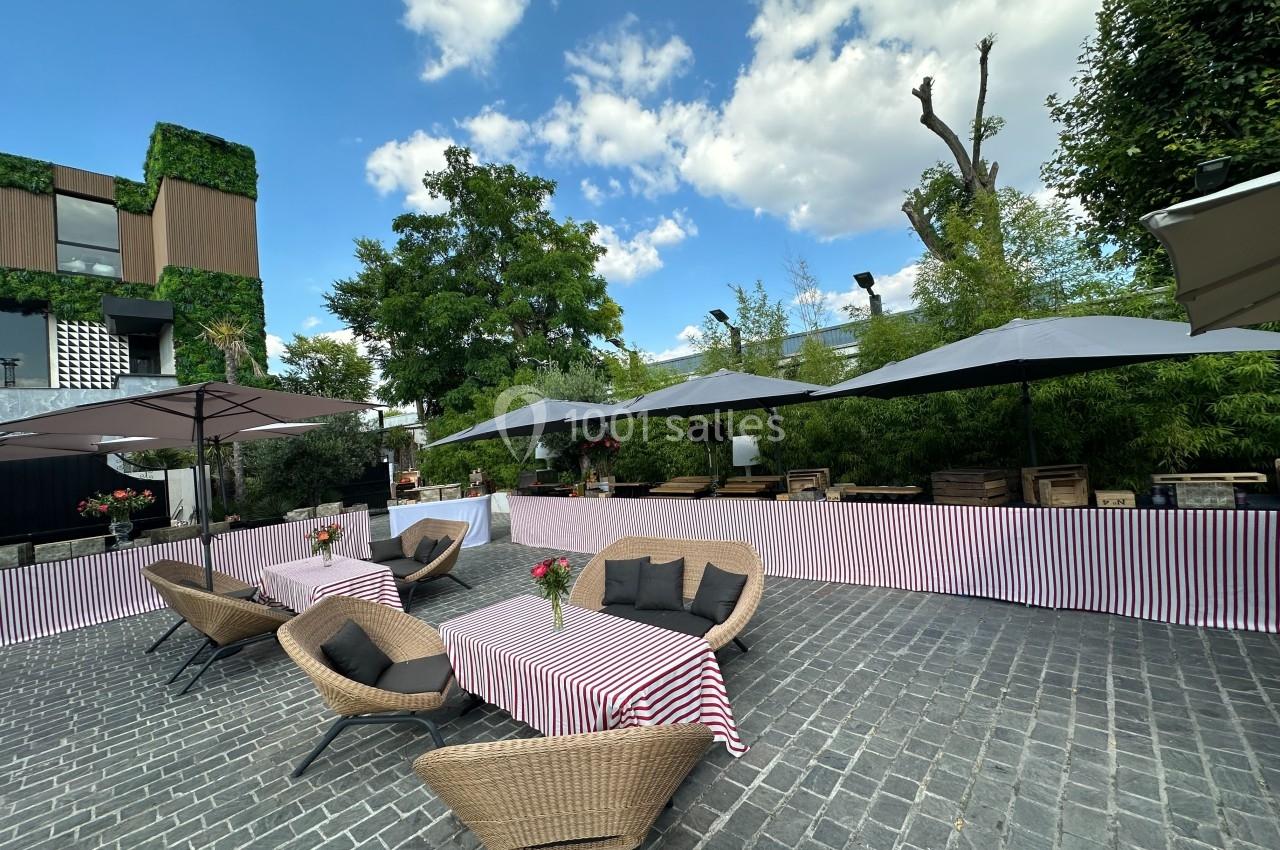 Terrasse extérieure avec tables couvertes de nappes rayées, chaises en osier et parasols sous un ciel partiellement nuageux.