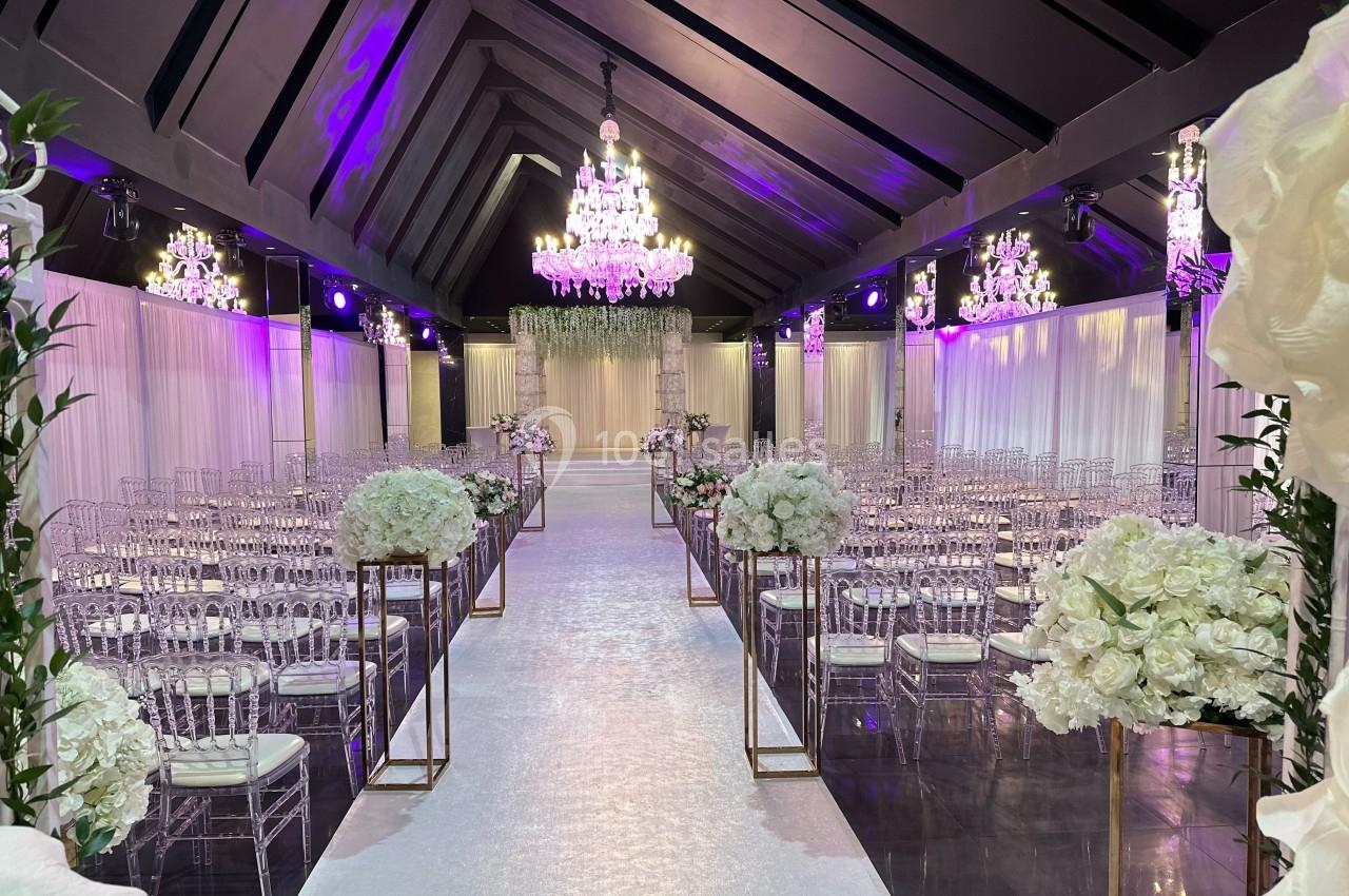 Salle de réception décorée pour un mariage, avec chaises transparentes, fleurs blanches et lustres suspendus.