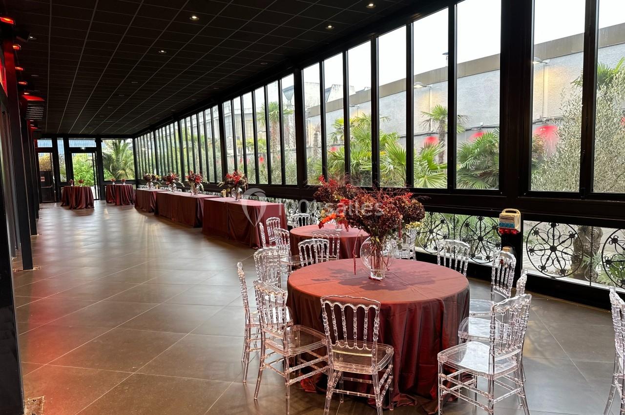 Salle lumineuse avec grandes baies vitrées, tables rondes dressées avec nappes rouges et chaises transparentes.