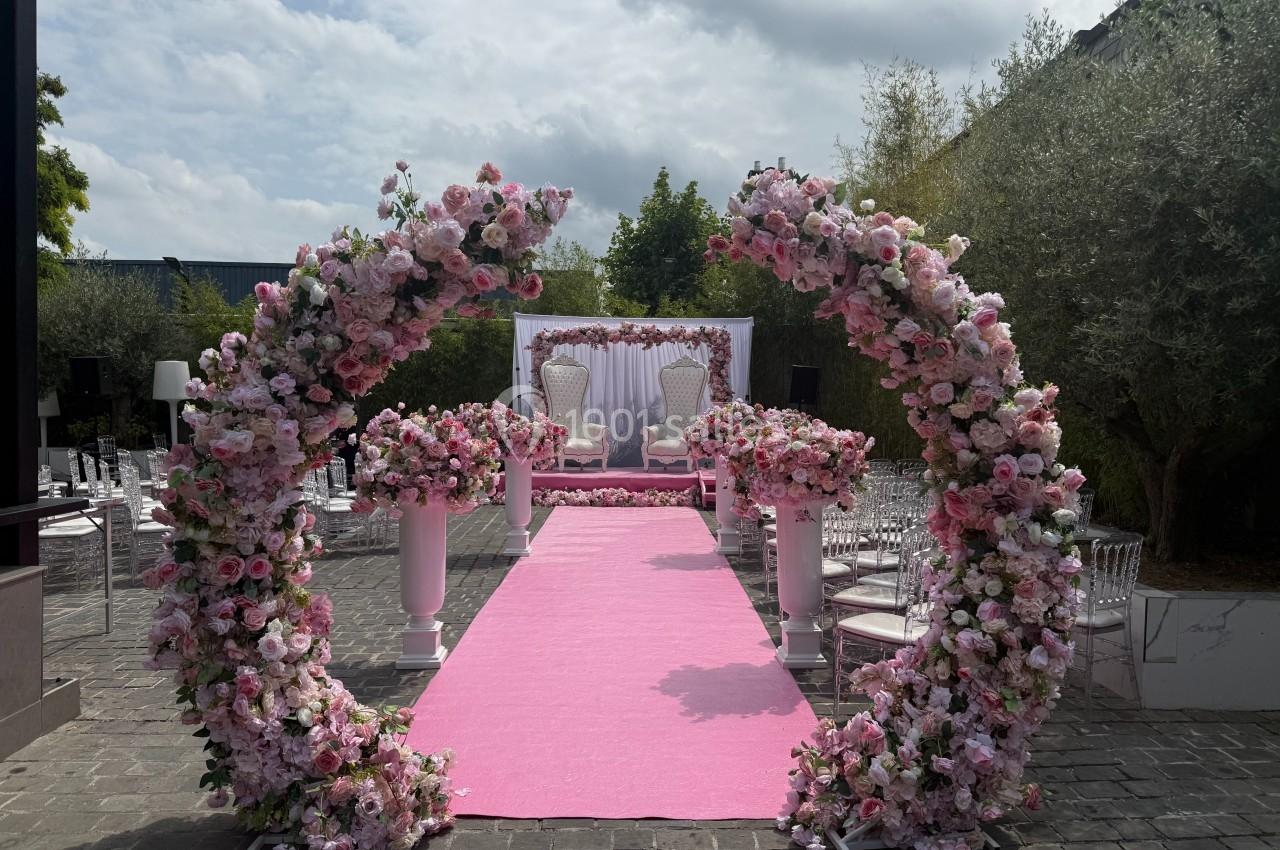 Allée centrale décorée de fleurs roses menant à une arche florale et deux fauteuils devant un fond blanc.
