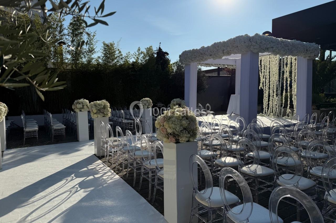 Allée de cérémonie de mariage en extérieur, décorée de fleurs blanches, avec chaises transparentes et arche florale.