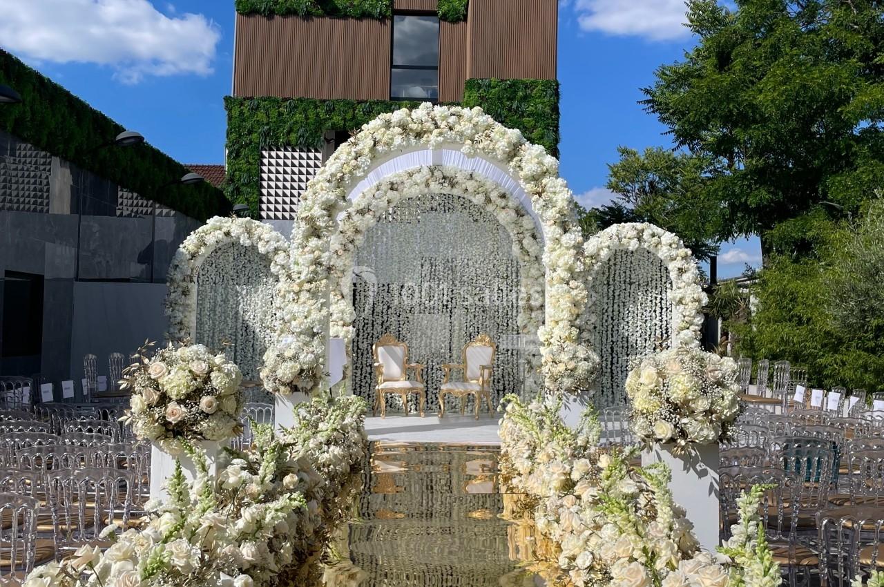 Allée décorée de fleurs blanches menant à une arche florale et deux chaises, avec un bâtiment moderne en arrière-plan.