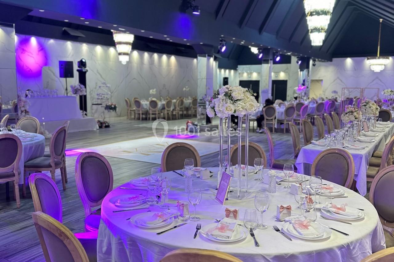 Salle de réception élégante avec tables rondes dressées, chaises en bois, éclairage violet et décorations florales.