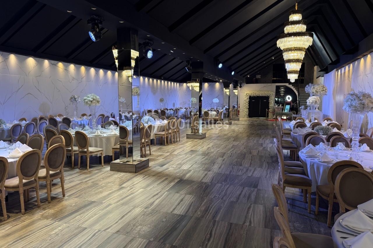 Salle de réception élégante avec tables dressées, chaises en bois clair, lustres imposants et éclairage tamisé.