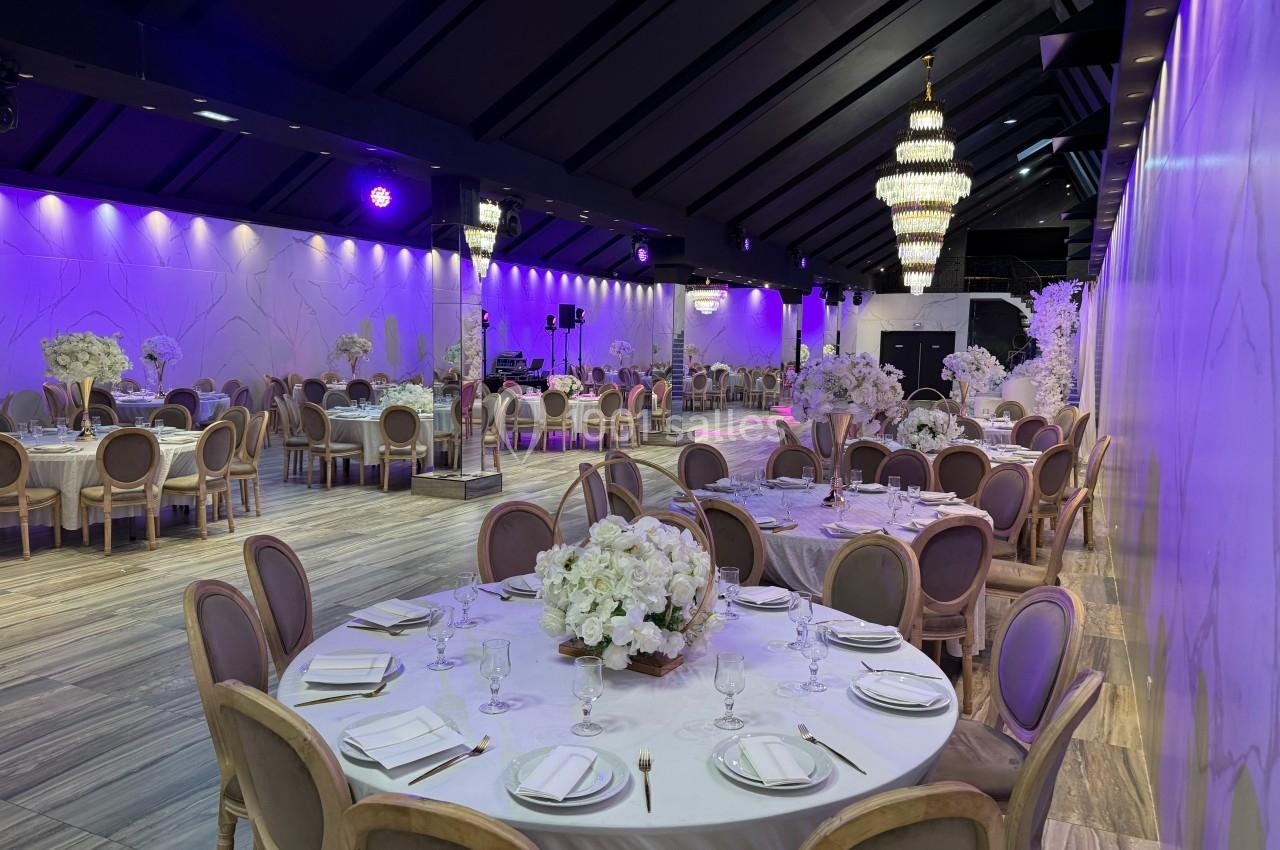 Salle de réception élégante avec tables rondes dressées, décorations florales blanches et éclairage violet tamisé.