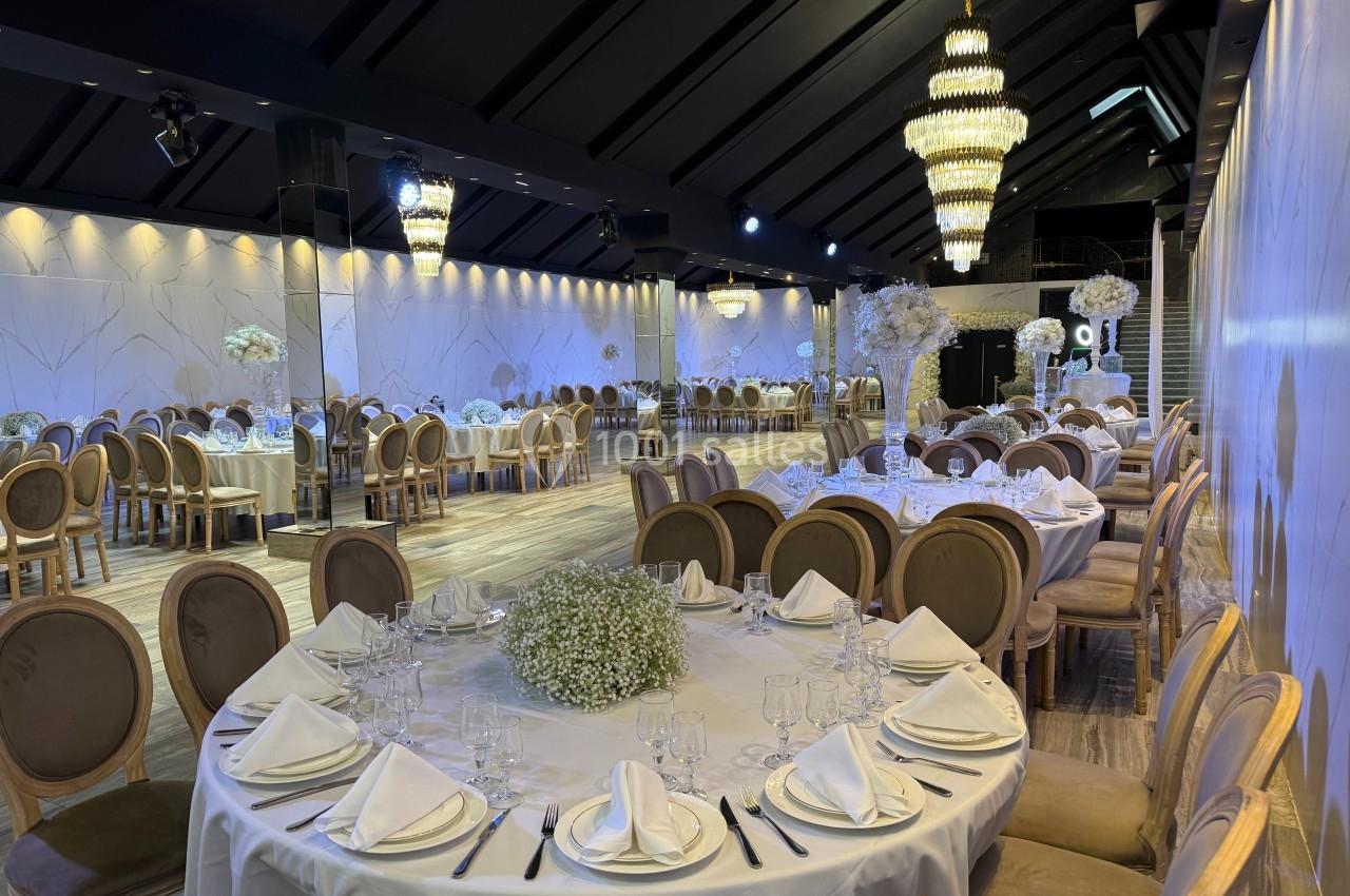 Salle de réception élégante avec tables rondes dressées, chaises en bois, décor floral blanc et grands lustres.
