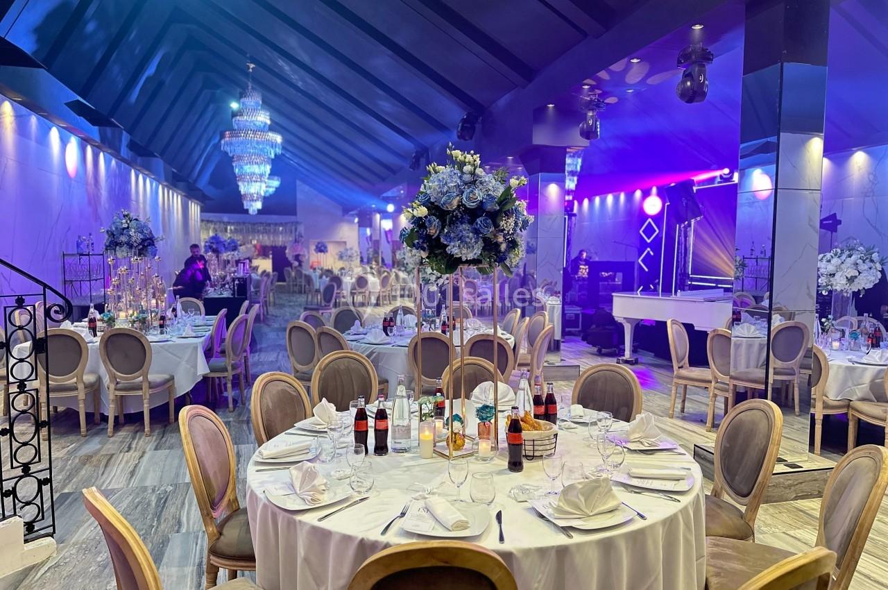 Salle de réception élégante avec tables dressées, chaises en bois, décor floral et éclairage tamisé.