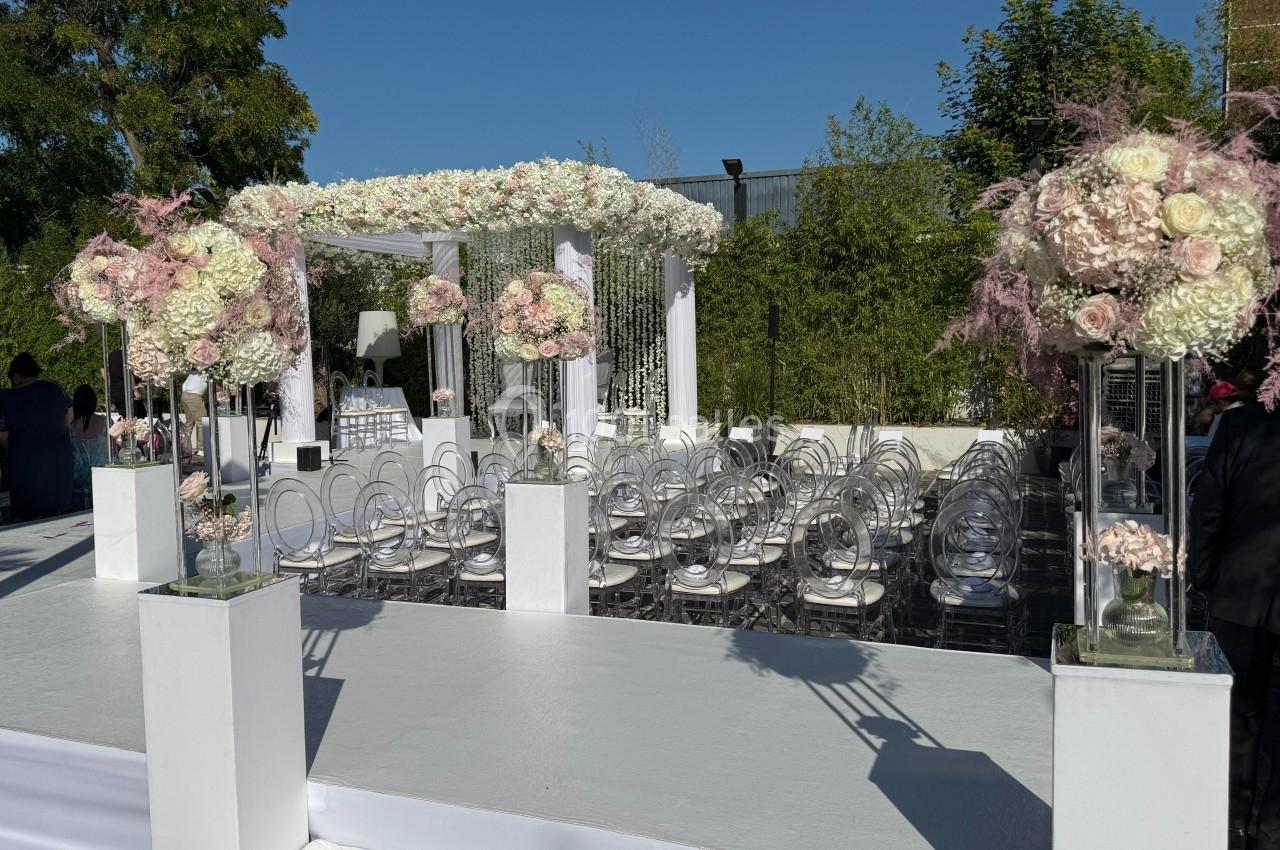 Structure de cérémonie en plein air décorée de fleurs blanches et roses, avec des chaises transparentes alignées.
