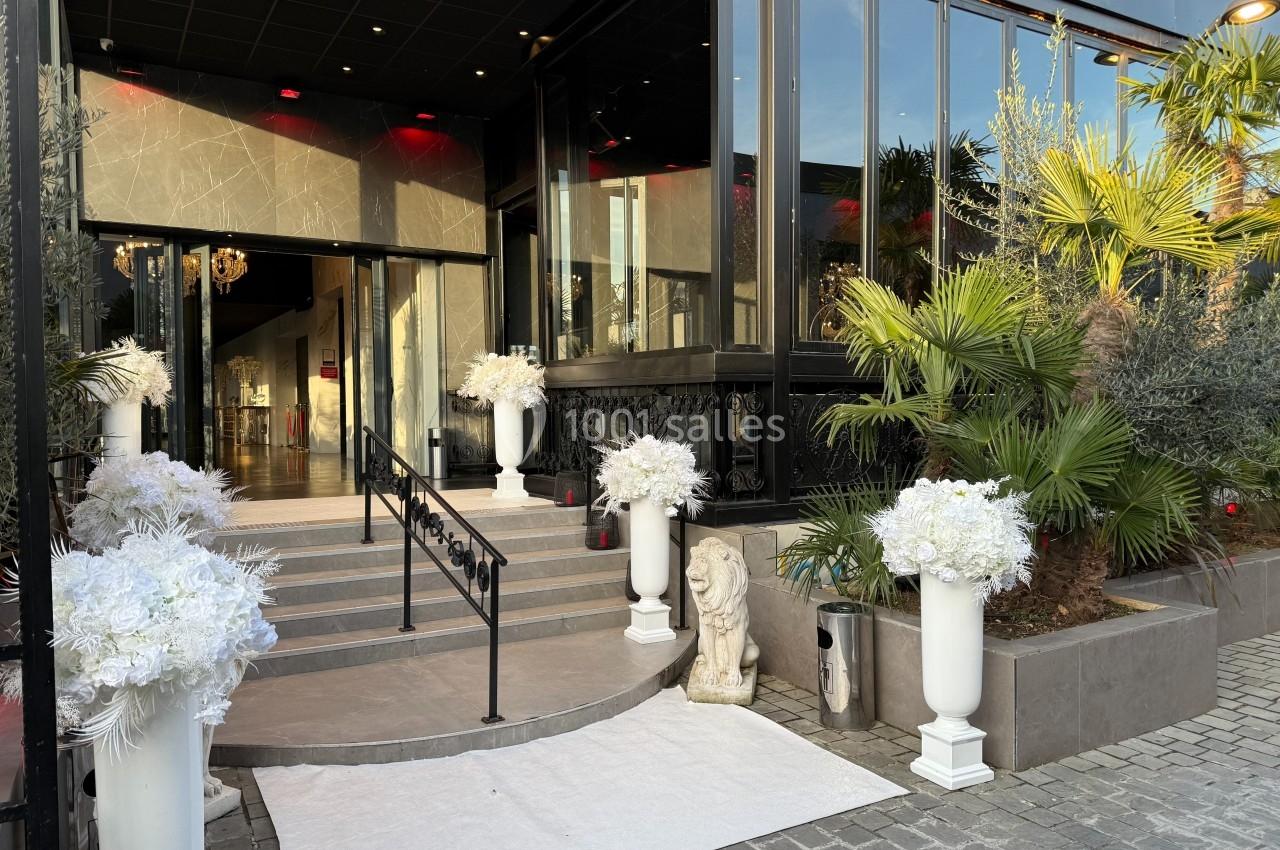 Entrée d'un bâtiment élégant avec des colonnes blanches décorées de fleurs, entourée de palmiers et d'une statue de lion.