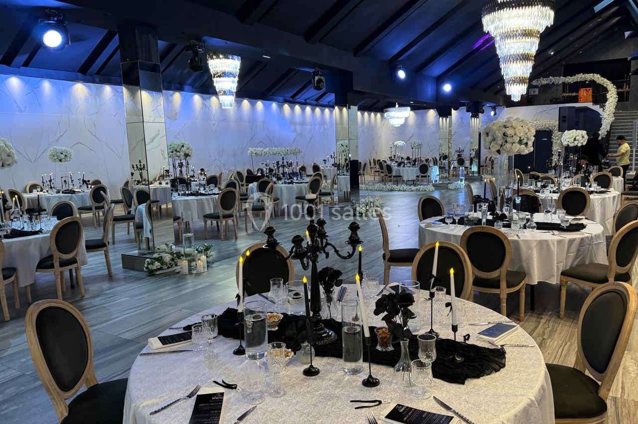 Salle de réception élégante avec tables dressées, chandeliers noirs, décorations florales blanches et éclairage tamisé.